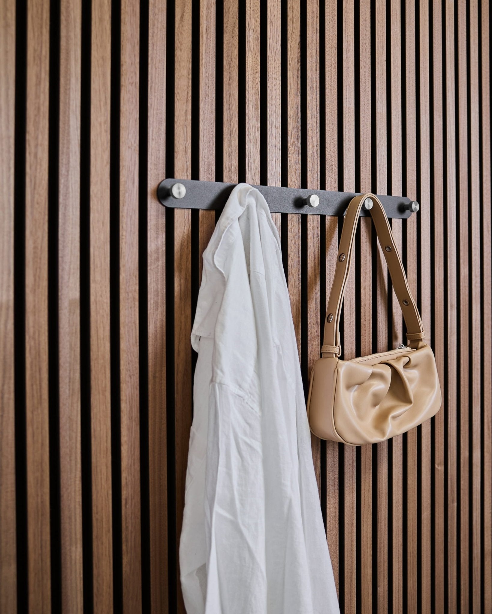 A white shirt and beige handbag hang on WoodUpp Norr X5 Black/Stainless Steel coat hooks attached to a vertical acoustic slatwall panel.